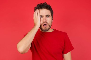 Utanmış, genç, yakışıklı, beyaz bir adam kırmızı arka plan üzerinde tişört giyiyor, şok olmuş bir ifadeyle, şaşkın bir şekilde içeride poz veriyor.