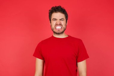 İzole edilmiş kırmızı arka plan üzerinde tişört giyen kızgın, genç, beyaz bir adamın portresi dişlerini sıkar, tatminsizlikten kaşlarını çatar, görevleri yüzünden tedirgin olur. Öfkeli adam pencereden dışarı bakar, sefil havayı görür.