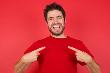 İzole edilmiş kırmızı arka plan beton noktalarının üzerine tişört giyen yakışıklı beyaz bir adam. Alışverişe gidip başarılı alışverişler yaptıktan sonra iyi bir ruh hali içinde.