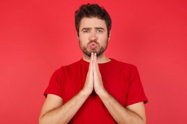 İzole edilmiş kırmızı arka planda tişört giyen yakışıklı beyaz adam mutlu bir şekilde gülümsüyor, soruşturmacıdan hoş haberler aldığına seviniyor, avuç içlerini bir arada tutuyor, insanların duygularını bir arada tutuyor..