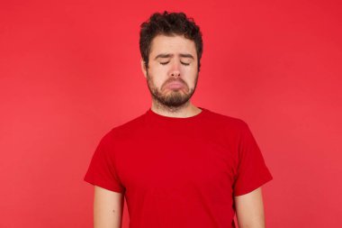 Kasvetli, reddedilmiş, kırmızı arka plan üzerinde tişört giyen yakışıklı beyaz adamın sorunları ve zorlukları var, alt dudak kıvrımları var ve umutsuzluk içinde gözlerini kapatıyor, umutsuz denemeler sonrasında depresyonda. 