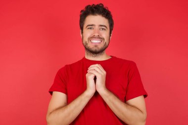 İzole edilmiş kırmızı arka planda tişört giyen yakışıklı beyaz adam mutlu bir şekilde gülümsüyor, soruşturmacıdan hoş haberler aldığına seviniyor, el ele tutuşuyor. İnsan duyguları kavramı.