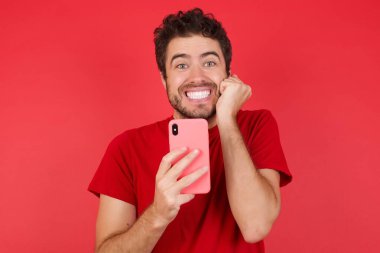 İzole edilmiş kırmızı arka plan üzerine tişört giyen neşeli genç beyaz adam cep telefonu cihazıyla poz veriyor, modern akıllı telefona mesaj yazıyor, boş zamanlarında komik videolar izliyor, iyi internet bağlantılarından hoşlanıyor., 
