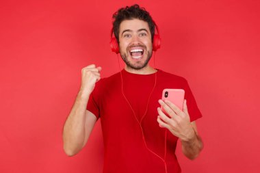 İzole edilmiş kırmızı arka plan üzerine tişört giyen genç, yakışıklı beyaz adam kulaklığa bağlı modern cep telefonunu tutuyor, iyi duygulardan dolayı yumruğunu sıkıyor, sevinçle haykırıyor., 