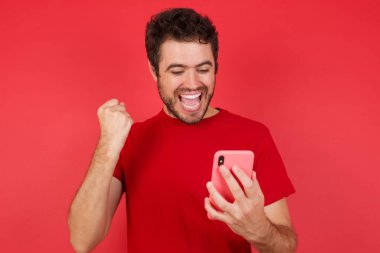 İzole edilmiş kırmızı arka plan üzerinde tişört giyen yakışıklı beyaz bir adam hücrede oturmuş kitap okuyor. 