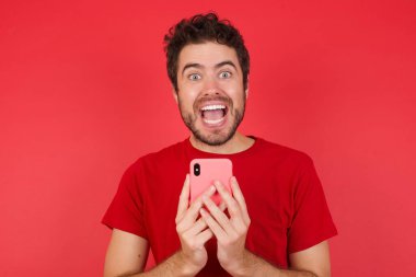 Fotokopi uzay fotoğrafında, genç yakışıklı beyaz adam izole edilmiş kırmızı arka plan üzerinde tişört giyiyor ve elinde telefon tutuyordu.