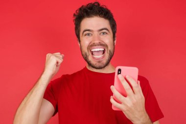 İzole edilmiş kırmızı arka plan üzerinde tişört giyen yakışıklı beyaz adamın portresi. Hücrede kutlama yapıyor. 