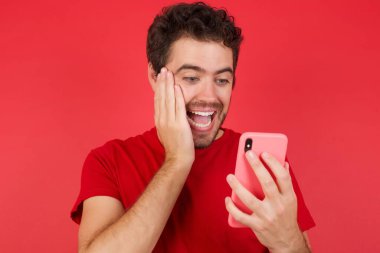 İzole edilmiş kırmızı arkaplan üzerine tişört giyen yakışıklı beyaz adamın yakın plan portresi. Yeni uygulama 5G kullanarak cep telefonunda SMS okuyor. 