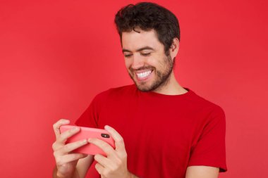 İzole edilmiş kırmızı arka plan üzerinde tişört giyen genç beyaz adamın yakın plan portresi. Hücrede el ele tutuşmuş video oyunları oynuyor ya da sohbet ediyor. 