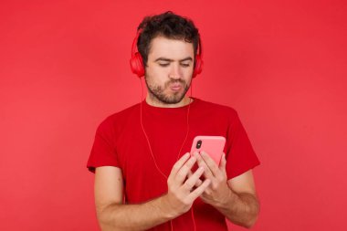 Deli gibi etkilenmiş genç beyaz bir adam kırmızı arka plan üzerinde tişört giyiyor cep telefonu kullanıyor sosyal medyada inanılmaz haberler okuyor, haykırıyor vay canına, şık kıyafetler giyiyor.