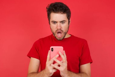 İzole edilmiş kırmızı arka plan üzerinde tişört giyen yakışıklı beyaz adamın portresi sosyal medya haberleri ya da önemli e-postalar okuyarak akıllı telefon kullan 