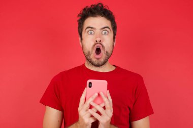 Şok olmuş genç, yakışıklı, beyaz bir adam izole edilmiş kırmızı arka plan tişörtü giyiyor. Ağzını aç telefon aç. Reklam ver. İnanılmaz büyük alışveriş fiyatları. 