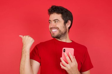 Kırmızı arka plan üzerinde tişört giyen yakışıklı beyaz bir adam akıllı telefonuyla mesaj atıyor ve gülümseyen yüzüyle baş parmağıyla onu işaret ediyor.