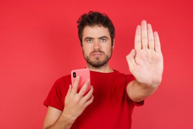 Yakışıklı, beyaz bir adam kırmızı arka plan üzerinde tişört giyiyor. Akıllı telefonuyla mesajlaşıp duruyor. Ciddi ve kendinden emin bir ifadeyle dur işareti yapıyor.