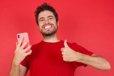 Kırmızı arka plan üzerinde tişört giyen yakışıklı beyaz adam akıllı telefonuyla mesajlaşıyor ve kullanıyor. Gülümsüyor. Tamam işareti yapıyor, başparmağını parmaklarıyla kaldırıyor.