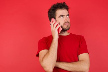 Üzgün, genç, yakışıklı, beyaz tişörtlü, izole edilmiş kırmızı arka planda akıllı telefondan konuşan bir adam. İletişim konsepti.