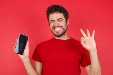 İzole edilmiş kırmızı arka plan üzerinde tişört giyen yakışıklı beyaz bir adam hücrede el ele tutuşmuş, tamam işareti gösteriyor. 