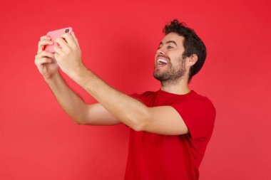 Genç, beyaz bir adam, izole edilmiş kırmızı arka plan tişörtü giyiyor. Sosyal medyaya göndermek için selfie çekiyor. Ya da arkadaşlarıyla görüntülü görüşme yapıyor..