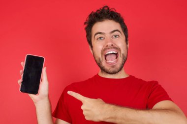 İzole edilmiş kırmızı arka plan üzerinde tişört giyen yakışıklı beyaz adam siyah ekranı gösteren hücrede el ele tutuşuyor. 