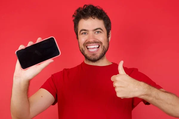 年轻英俊的高加索男子穿着T恤衫 罩在孤立的红色背景上 手捧着显示黑色屏幕大拇指的手机 — 图库照片
