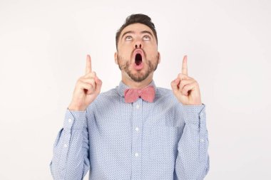 Beyaz stüdyodaki beyaz adam, papyon takıyor, şaşırmış ve şaşırmış görünüyor ve parmaklarıyla işaret ediyor ve kollarını kaldırıyor.
