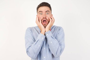 Beyaz stüdyodaki beyaz adam, papyon takıyor, heyecan ve heyecan ifade ediyor, çenesi düşük, elleri yanaklarında, gözleri fırlamış