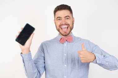 Beyaz stüdyodaki neşeli beyaz adam, papyon takıyor, cep telefonu cihazı tutuyor, akıllı telefon sunuyor, siyah ekran başparmağını kaldırıyor. 