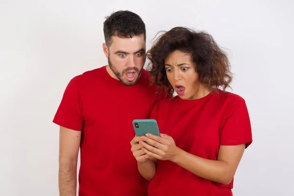 年轻貌美的年轻夫妇穿着白色背景的红色T恤 他们的画像用智能手机阅读社交媒体新闻或重要的电子邮件 — 图库照片