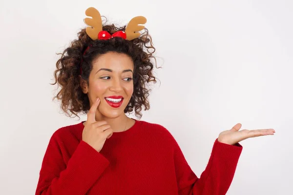 stock image woman in red sweater and deer horns headband posing in studio with smile, showing copy space wall