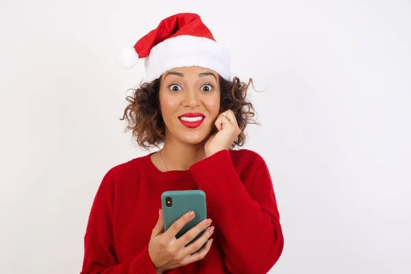 戴着圣诞老人帽子的女人在工作室里浏览手机 微笑着看着相机 — 图库照片