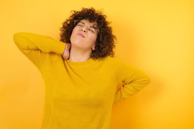 Stüdyoda kıvırcık saçlı, sarı kazaklı bir kadın boyun ağrısı çekiyor, eliyle boynuna dokunuyor, kas ağrısı çekiyor.