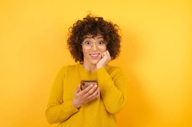  Stüdyoda kıvırcık saçlı, akıllı telefonlu, elleri yanağında bir kadın. 
