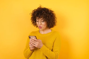 Stüdyoda sarı kazaklı, kıvırcık saçlı bir kadın, cep telefonunu karıştırıyor., 