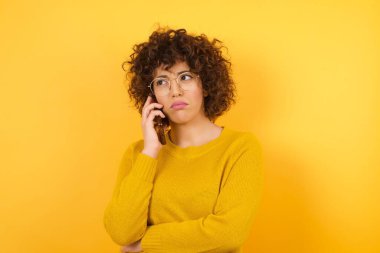 Stüdyoda saçları kıvırcık, sarı arka planı ve cep telefonu olan üzgün bir kadın. 