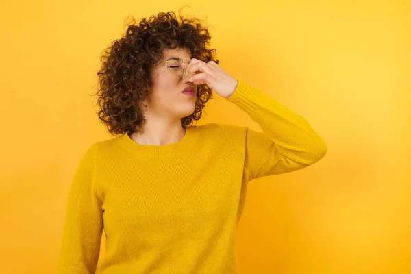 woman in yellow sweater with curly hair in studio, smelling something stinky and disgusting, intolerable smell, holding breath with fingers on nose. Bad smell