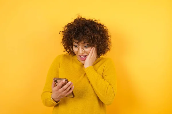 surprised woman with curly hair in yellow studio, holding and browsing mobile phone