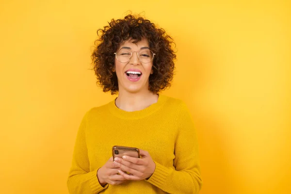 happy woman with curly hair in studio with smartphone, eye wink on camera