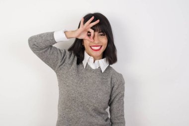 Genç beyaz kadın el hareketleriyle gülümsüyor, mutlu yüzüyle parmaklarının arasından bakıyor..