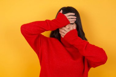 Sarı arka planda genç bir kadın. Elleriyle gözlerini ve ağzını kapatmış, şaşırmış ve şok olmuş. Duyguları gizlemek.