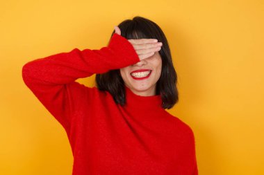 İzole edilmiş sarı arka planda duran genç beyaz bir kadın. Gülümsüyor ve gülümsüyor. Gözleri şaşkınlıkla örtüyor. Kör kavram.