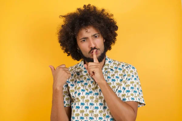 Young handsome man  asking to be quiet with finger on lips pointing with hand to the side. Silence and secret concept.