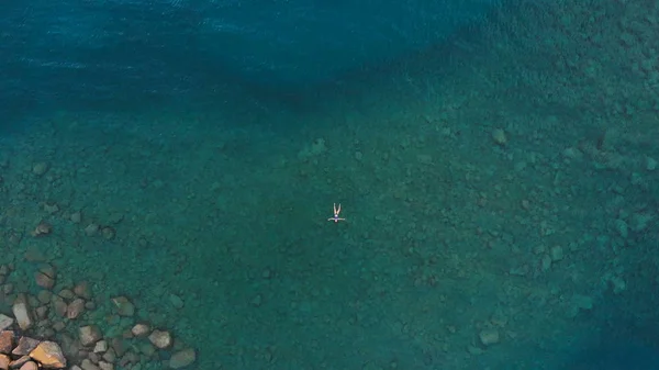 AERIAL: woman floating on blue water surface, swimming in transparent mediterranean sea, top down view, summer vacation concept