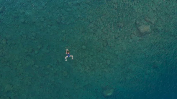 AERIAL: woman floating on blue water surface, swimming in transparent mediterranean sea, top down view, summer vacation concept