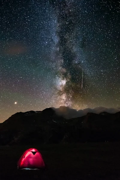 Shooting star with green tail starry sky milky way. Illuminated tent in the foreground, camping on the Alps. Mars Planet on the left. Adventure and exploration in summertime.