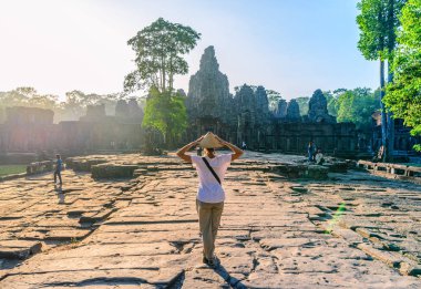 Bir turist ziyaret Bayon Tapınağı, Angkor Thom kalıntıları, hedef Kamboçya seyahat. Geleneksel şapka ve yükseltilmiş kollar, kadın ile arka görünümü, gündoğumu sunburst arka ışık.