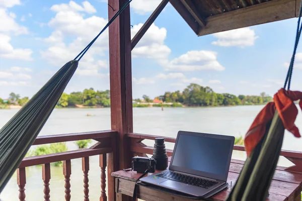 Trabalhando com laptop em varanda de madeira em resort turístico com rede pendurada no Rio Mekong, Laos. Conceito de milênios trabalhando em todo o mundo e nômades digitais . Imagens De Bancos De Imagens