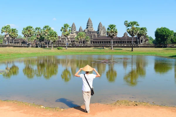 Un turista que visita las ruinas de Angkor Wat al amanecer, destino de viaje Camboya. Mujer con sombrero tradicional y brazos levantados, vista trasera, reflexión fachada principal en estanque de agua — Foto de Stock