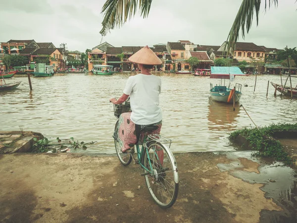 Woman with traditional vietnamese hat cycling on the river bank at Hoi An, famous travel destination in Vietnam.  River flooding in the street monsoon season. — Stock Photo, Image