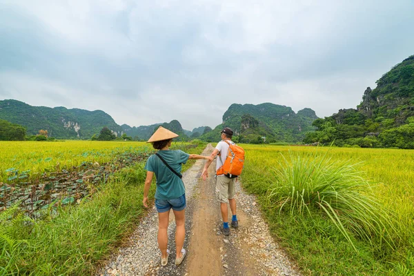 Pareja caminando mano en el camino del campo entre campos de arroz. Hombre con mochila y mujer con sombrero vietnamita divirtiéndose viajando juntos de vacaciones. Tam Coc Trang An Ninh Binh karst rock mountain ran — Foto de Stock