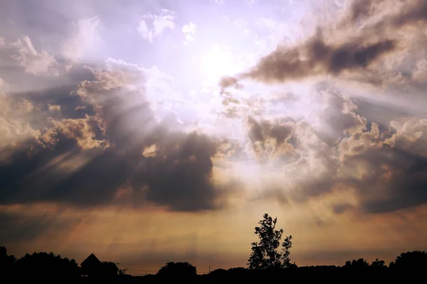 Cloudy Sunset over the Village. The Rays of the Sun Shine Through the Clouds. On the Horizon Silhouette of Trees and the Roof of the House.